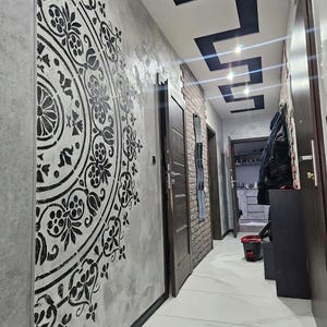 Op de afbeelding: Een gang met een decoratieve muur met een groot zwart bloemenontwerp op een grijze, getextureerde achtergrond. Het plafond heeft zwarte geometrische accenten en de vloer is wit betegeld. Verschillende bruine deuren en een bakstenen muur zijn zichtbaar.