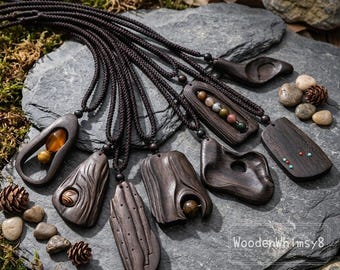 May include: A collection of dark brown wooden pendant necklaces with various carved designs. The pendants are suspended on black cords and rest on a gray stone surface. Some pendants feature inlaid stones or unique shapes.