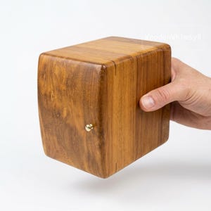 以下が含まれることがあります： 温かみのある茶色の仕上げと小さな金色のノブが付いた木製の箱。 箱は丸みを帯びたエッジと滑らかな表面を持ち、天然の木目を際立たせています。 箱は手で持たれています。