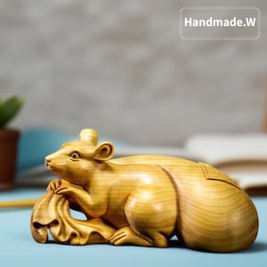 Puede incluir: Escultura de madera de una rata, tallada en madera de color claro. La rata se representa en posición reclinada, sosteniendo un objeto estilizado en forma de hoja. La escultura tiene un acabado liso y pulido, con el texto "Handmade.W".