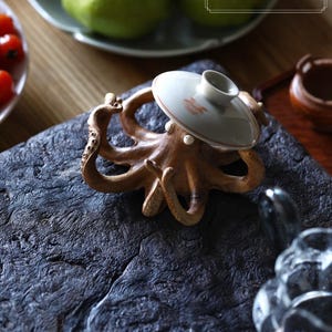 May include: A brown octopus-shaped ceramic sculpture with a white lid, resting on a dark, textured surface. The lid has a red and orange floral design. The sculpture is a decorative item.