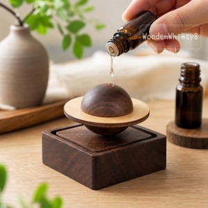 May include: A dark brown wooden essential oil diffuser in the shape of a planet with a light brown ring. A small brown glass bottle is pouring oil onto the diffuser. The diffuser sits on a dark brown square base.