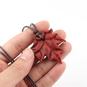 以下が含まれることがあります： 彫刻された木製のカエデの葉のペンダントネックレス。ペンダントは濃い赤茶色で、茶色のコードで吊り下げられています。葉のデザインには詳細な葉脈があり、自然な仕上がりです。