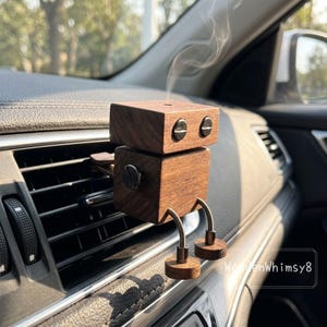 May include: A wooden robot-shaped car air freshener. The robot has a two-tiered head and body, with silver accents for eyes and a screw. It has metal legs and circular feet. Smoke emanates from the top, suggesting it is in use. The air freshener is placed on a car vent.