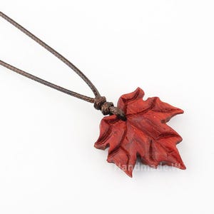 Può includere: Collana con ciondolo a forma di foglia d'acero in legno marrone-rossastro fatto a mano. La foglia è intagliata in modo intricato con venature dettagliate e pende da un cordoncino marrone. La collana è esposta su sfondo bianco.
