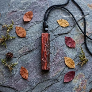 Op de afbeelding: Een handgemaakte houten hangerketting met een getextureerde, roodbruine rechthoekige hanger. De hanger hangt aan een zwart koord en is omgeven door herfstbladeren en mos op een grijze stenen ondergrond. De tekst "WoodenWhimsy8" is zichtbaar.