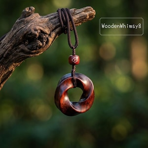 Peut inclure: Un collier pendentif en bois fait à la main avec un cordon marron foncé. Le pendentif est une forme d'anneau torsadé brun rougeâtre foncé. Une petite perle rouge avec un motif de tourbillon blanc est au-dessus du pendentif. Le texte "WoodenWhimsy8" est dans le coin supérieur droit.