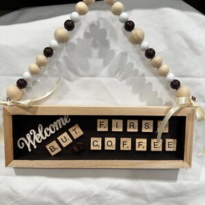 以下が含まれることがあります： 「Welcome But First Coffee」と文字タイルで書かれた木製の看板。看板は茶色と白のビーズのガーランドから吊り下げられています。