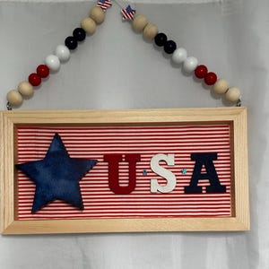 以下が含まれることがあります： 赤と白のストライプの生地が内側に敷き詰められた木製の額縁付きの看板。生地には青い星と「USA」の文字が赤、白、青の文字で書かれています。看板は、赤、白、青のアクセントが付いた木製のビーズの紐から吊り下げられています。