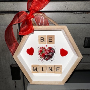 以下が含まれることがあります： 六角形の木製フレームに入ったバレンタインデーの装飾。白い内側には、Scrabbleのタイルで綴られた「BE MINE」の文字、2つの赤いハート、花のハートのデザインが施されています。上部には赤いリボンが結ばれています。