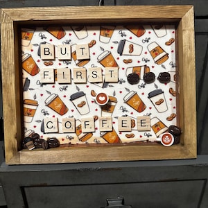 以下が含まれることがあります： 木製のフレームに入ったコーヒーをテーマにしたアートワーク。背景にはコーヒーカップと豆の繰り返し模様があります。「BUT FIRST COFFEE」というフレーズは、スクラブルのタイルを使って綴られています。コーヒー豆とコーヒーカップがアートワークを飾っています。