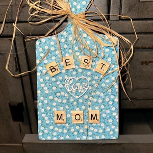 以下が含まれることがあります： 花柄の青いまな板で、木製の文字タイルで「BEST MOM」と書かれています。中央には「love」という言葉が入った白いハートがあります。ボードはラフィアで飾られています。