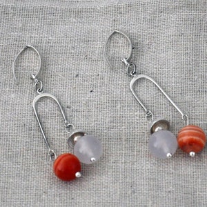 Könnte beinhalten: Ein Paar silberne Ohrringe mit einem einzigartigen Design. Jeder Ohrring hat einen silbernen Bogen, an dem ein weißer und ein roter Edelstein hängen.