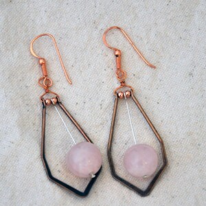May include: Art Deco earrings with rose quartz and copper. The earrings have a geometric design with a teardrop shape, a light pink quartz bead, and a copper hook. The earrings are made of copper and quartz.
