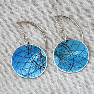 May include: A pair of silver earrings. Each earring has a circular blue design with black abstract lines and a curved silver hook. The text "Atomic sterling silver earrings" is on the left side of the image.