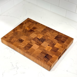 May include: A rectangular end-grain butcher block cutting board. The board is made of various wood pieces, creating a checkerboard pattern. It has a warm, natural wood tone with visible grain and a handle cut out on one side.