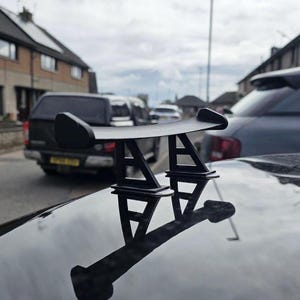May include: A black car spoiler, mounted on the trunk of a vehicle. The spoiler has a curved wing design supported by two angled brackets. The image is taken outdoors with other vehicles in the background.