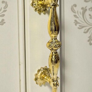 May include: An ornate, gold-coloured cabinet pull with a detailed floral design. The pull is mounted on a white cabinet door with a subtle, grey floral pattern. The hardware features a decorative, vertical handle.