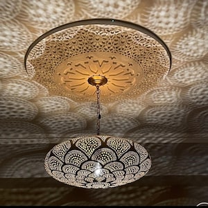 Puede incluir: Una lámpara colgante ovalada con una pantalla de metal calado y ornamentado. Está suspendida de una roseta de techo decorativa, proyectando sombras intrincadas. El diseño general tiene una estética cálida y acogedora.