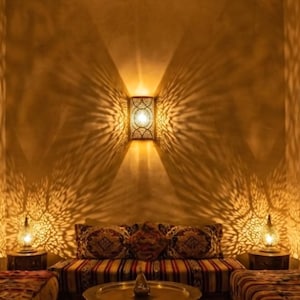 Peut inclure: Une applique murale rectangulaire avec un motif découpé complexe, projetant des rayons de lumière symétriques sur le mur. Deux lampes assorties plus petites sont posées sur des tables, éclairant une table ronde basse et des coussins.