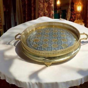 Op de afbeelding: Een ronde, gouden serveerschaal met een decoratieve, blauw-gouden gepatenteerde bodem. De schaal heeft een verhoogde rand en drie poten.