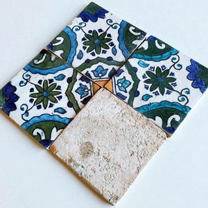 Könnte beinhalten: Anordnung von dekorativen Keramikfliesen in Rautenform. Die Fliesen zeigen florale Muster in Blau, Grün und Weiß. Eine Fliese hat eine strukturierte, beige Oberfläche, die sich von den gemusterten Fliesen abhebt. Für Inneneinrichtung.