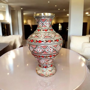 Könnte beinhalten: Verzierte Metallvase mit silbernem Finish, mit aufwendigen roten und weißen geometrischen Mustern. Die Vase hat einen breiten Sockel, einen bauchigen Körper und eine ausgestellte Oberseite. Die Vase steht auf einem hellrosa Tisch.