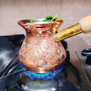 May include: A copper coffee pot with a brass handle and wooden grip sits on a gas stove. The pot is filled with green herbs and is heated by a blue flame. The pot has an embossed floral design.