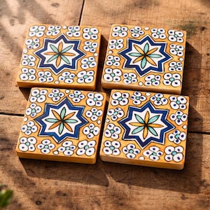 May include: Four square ceramic coasters with a repeating floral pattern and a central star design. The coasters feature a yellow background with blue, white, and orange accents. Each coaster measures approximately 4 inches by 4 inches.