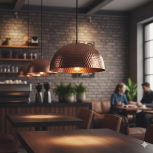 May include: Three copper-colored pendant lights with a hammered texture hang above wooden tables in a cafe. The lights are dome-shaped and have exposed light bulbs. The background features a brick wall, shelves, and a coffee machine.