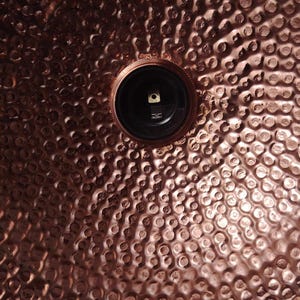 May include: Close-up of a copper-colored metal surface with a hammered texture. A black circular fixture is centered in the image. The surface has a dimpled pattern, creating a textured appearance.