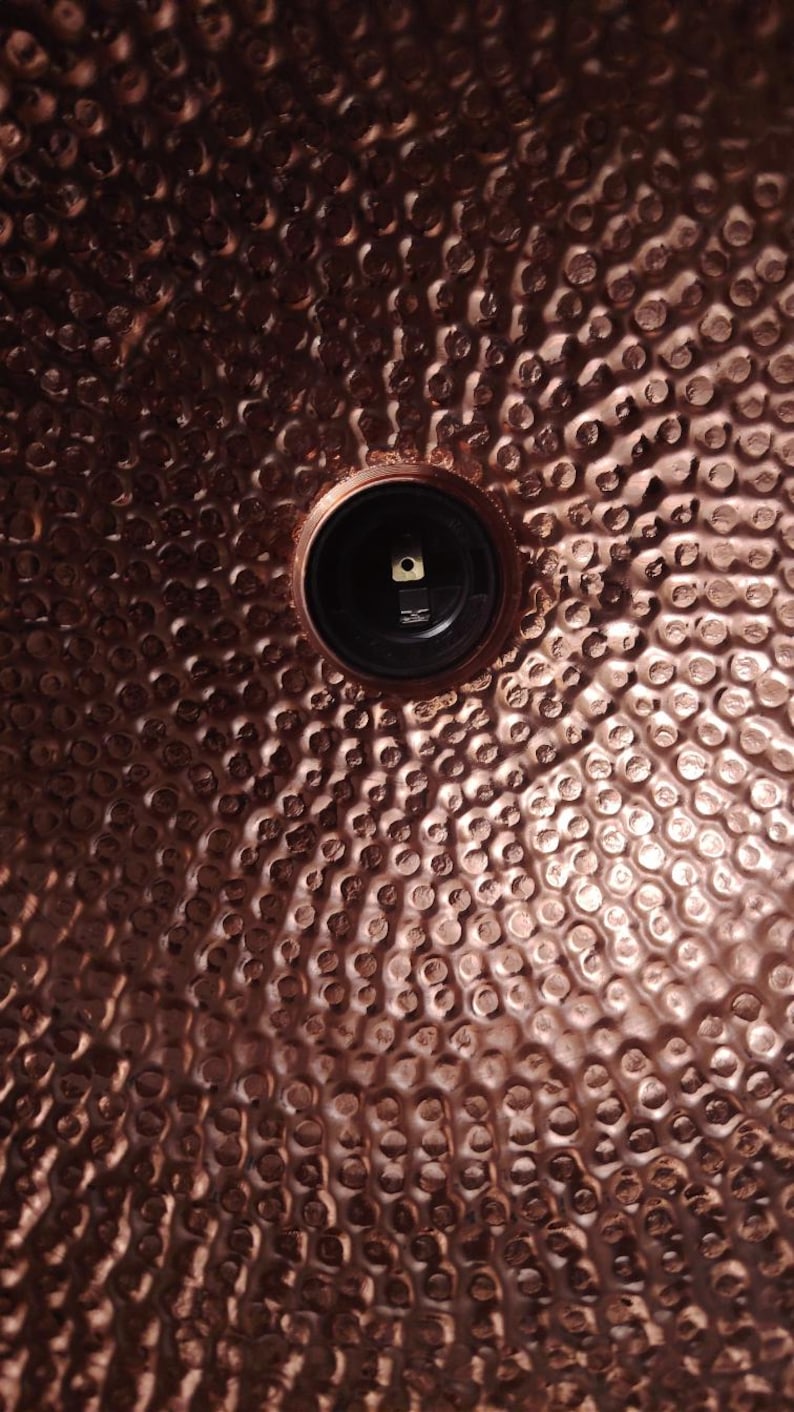 May include: Close-up of a copper-colored metal surface with a hammered texture. A black circular fixture is centered in the image. The surface has a dimpled pattern, creating a textured appearance.