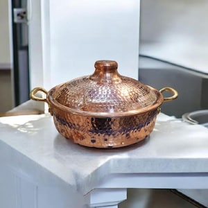 May include: A copper cooking pot with a lid, featuring a hammered texture. The pot has two handles and a knob on the lid. The pot is sitting on a white surface.