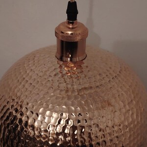May include: Copper pendant light with a hammered texture. The light fixture has a dome-shaped shade and a copper-colored top with a black cord. The light is designed to hang from the ceiling and provides ambient lighting.
