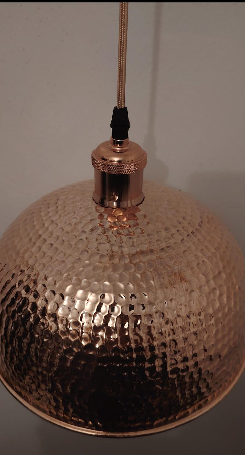 May include: Copper pendant light with a hammered texture. The light fixture has a dome-shaped shade and a copper-colored top with a black cord. The light is designed to hang from the ceiling and provides ambient lighting.