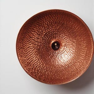 May include: A copper-colored, hammered metal bowl with a textured surface. The bowl has a rounded shape and a small, dark center. The bowl is likely used for decorative or functional purposes.