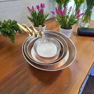 Puede incluir: Un juego de cuatro sartenes de cobre martillado con asas de latón, apiladas sobre una superficie de madera. Las sartenes varían en tamaño, con un interior brillante y reflectante. Flores moradas en macetas blancas y vegetación en el fondo.