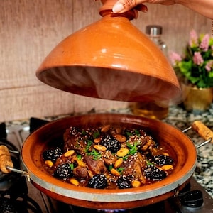 May include: A traditional Moroccan tagine dish, featuring a terracotta pot with a conical lid. The dish contains meat, plums, and almonds, garnished with herbs and sesame seeds. The tagine is on a gas stove, with steam rising from the dish.