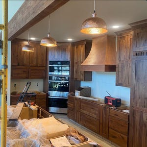 May include: A kitchen under construction with wooden cabinets and a copper range hood. Three copper pendant lights hang from the ceiling. The cabinets are a dark wood color, and the walls are white. A double oven is built into the cabinetry.
