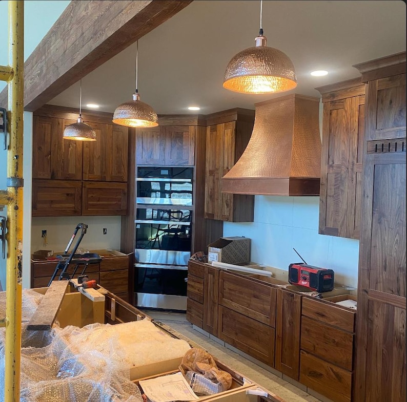 May include: A kitchen under construction with wooden cabinets and a copper range hood. Three copper pendant lights hang from the ceiling. The cabinets are a dark wood color, and the walls are white. A double oven is built into the cabinetry.