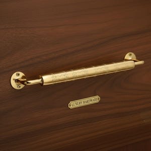 Könnte beinhalten: Ein goldfarbener Türgriff mit dekorativer Ätzung, montiert an einer dunkelbraunen Holztür. Der Griff ist mit "LUXURY HARDWARE" beschriftet.