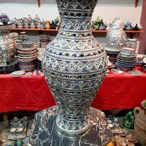 Könnte beinhalten: Eine große, verzierte Silbervase mit komplizierten geometrischen Mustern und farbenfrohen Akzenten. Die Vase steht auf einem Marmorständer, der ihre detaillierte Handwerkskunst zeigt. Weitere dekorative Gegenstände sind im Hintergrund zu sehen.