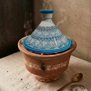 May include: A traditional terracotta tagine pot with a conical lid, adorned with a blue and white geometric pattern. Steam rises from the pot, suggesting it is in use. A gold spoon and small bowl of salt are nearby.