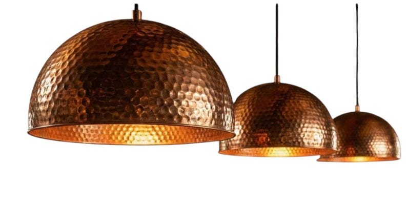 May include: Three copper pendant lights with a hammered texture and a warm, golden interior. The dome-shaped shades hang from black cords, creating a modern and stylish lighting fixture. The copper finish adds a touch of elegance.