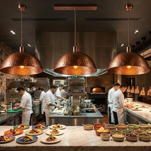 May include: A restaurant kitchen with three copper pendant lights hanging above the cooking area. Chefs in white coats are preparing food. Plates of food are arranged on a marble countertop. Copper pots hang on a brick wall.