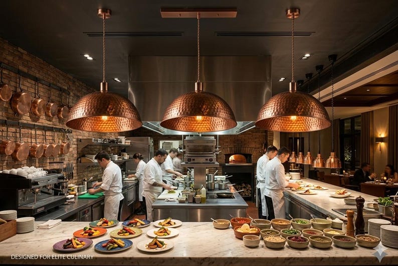 May include: A restaurant kitchen with three copper pendant lights hanging above the cooking area. Chefs in white coats are preparing food. Plates of food are arranged on a marble countertop. Copper pots hang on a brick wall.
