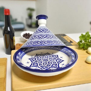 May include: A white and blue ceramic tagine pot with a conical lid. The pot features intricate blue patterns and a starburst design on the interior. The tagine is displayed on a wooden chopping board, with a bottle and other ingredients in the background.