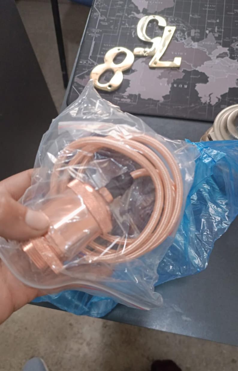 May include: A copper-colored pendant light fixture with a matching cord, packaged in a clear plastic bag. The fixture is a warm, metallic tone. Also visible are gold-colored house numbers.