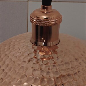 May include: Close-up of a copper pendant light fixture. The light has a textured, hammered metal shade and a copper-colored bulb socket. The cord is a woven, light brown color.