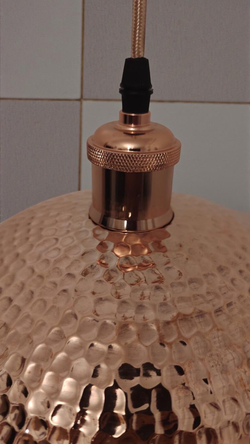 May include: Close-up of a copper pendant light fixture. The light has a textured, hammered metal shade and a copper-colored bulb socket. The cord is a woven, light brown color.